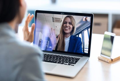Back view of female employee waving and speaking on video call with her colleague on online briefing with laptop at home.